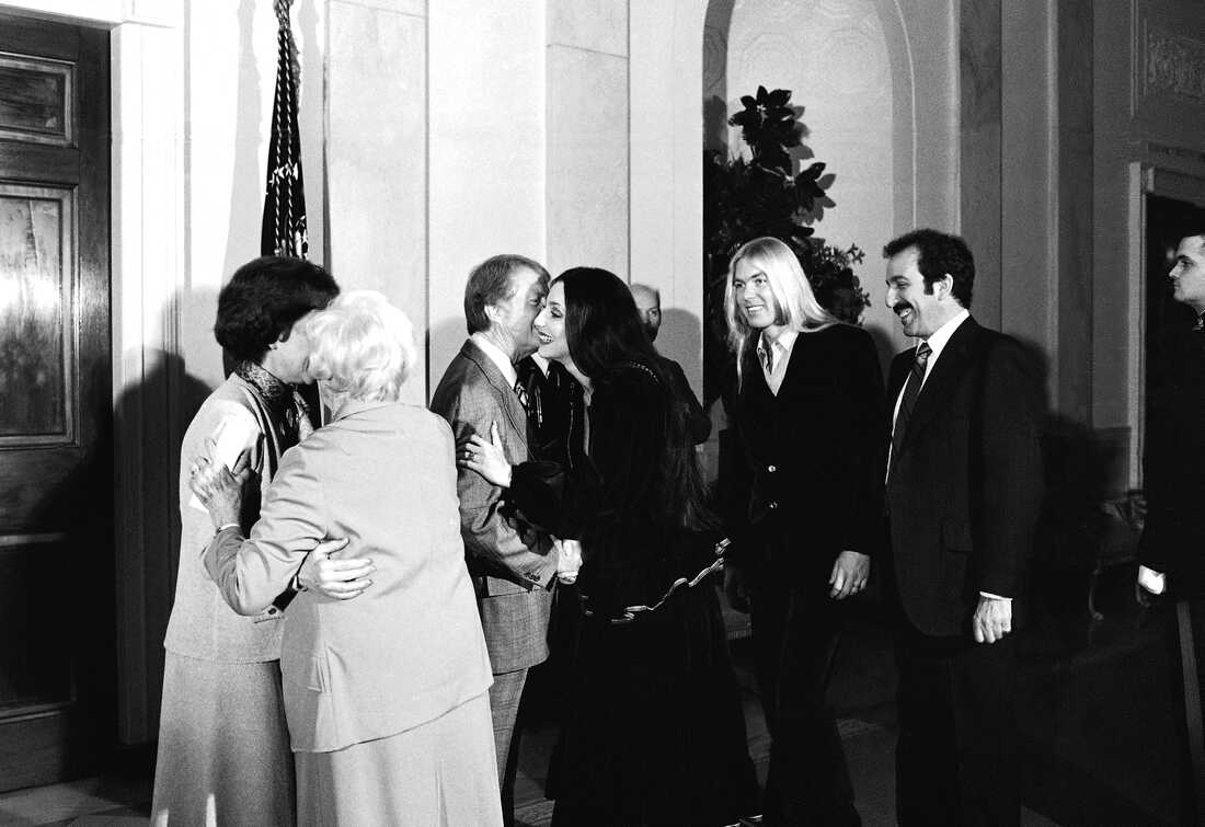 1977 年 1 月 21 日：总统吉米·卡特 (Jimmy Carter) 在白宫为花生旅 (Peanut Brigade)（一群竞选工作人员）举办招待会时，总统吉米·卡特 (Jimmy Carter) 亲吻了歌手雪儿 (Cher)，而她的丈夫格雷格·奥尔曼 (Gregg Allman) 站在一旁（右二）。