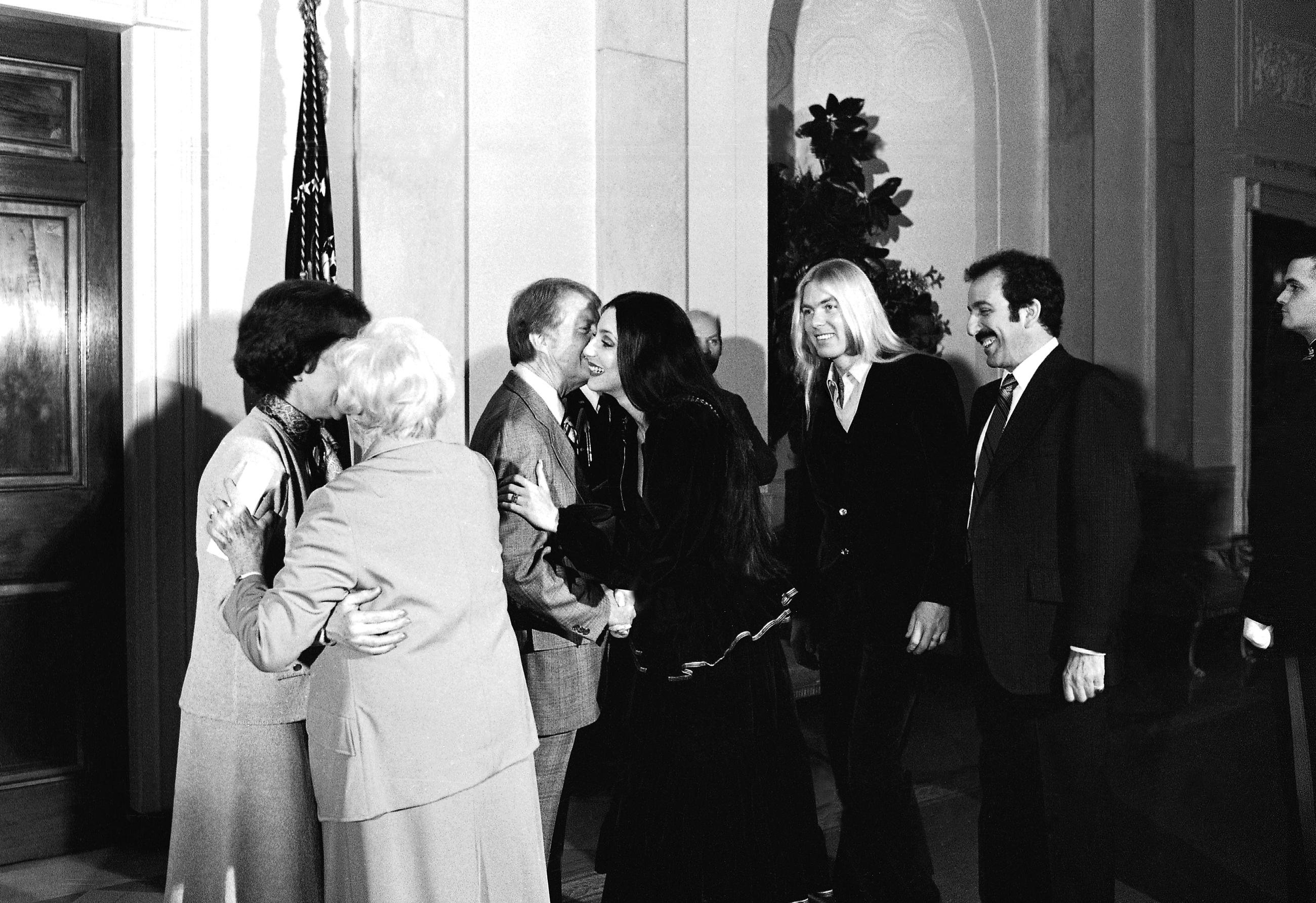 1977 年 1 月 21 日：总统吉米·卡特 (Jimmy Carter) 在白宫为花生旅 (Peanut Brigade)（一群竞选工作人员）举办招待会时，总统吉米·卡特 (Jimmy Carter) 亲吻了歌手雪儿 (Cher)，而她的丈夫格雷格·奥尔曼 (Gregg Allman) 站在一旁（右二）。