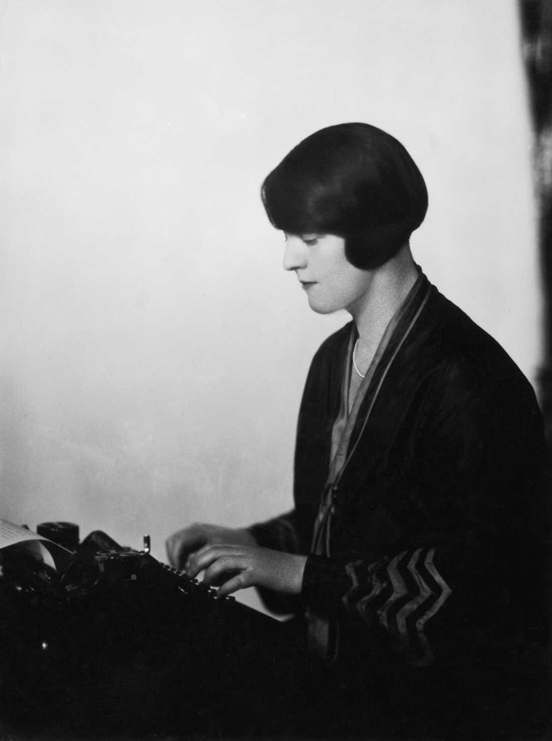 这张黑白照片显示了多萝西·汤普森(Dorothy Thompson)坐在打字机前,在钥匙上打字。她穿着深色连衣裙,短发短。