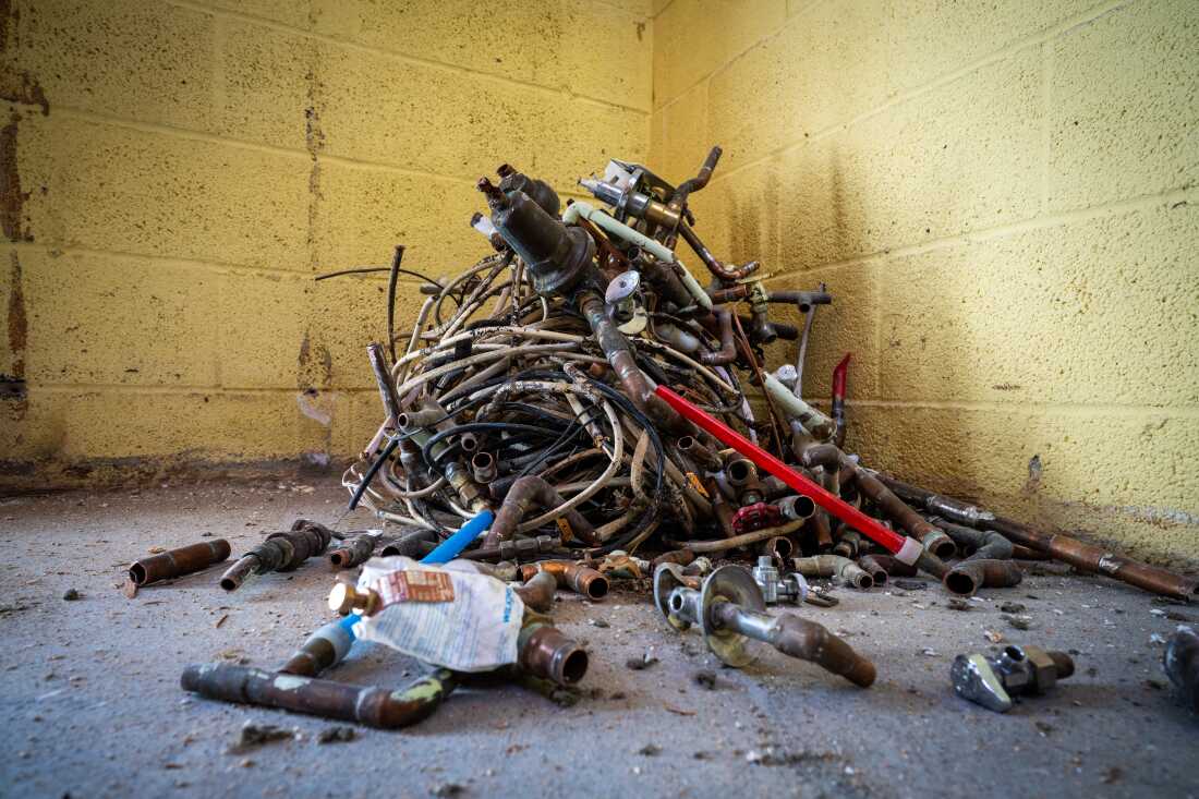 这张照片显示了一堆纠结的旧，厚的电线和金属管配件，位于家中地下室的角落。地下室有浅黄色的煤渣块墙壁和灰色混凝土地板，上面覆盖着灰尘和砂砾。