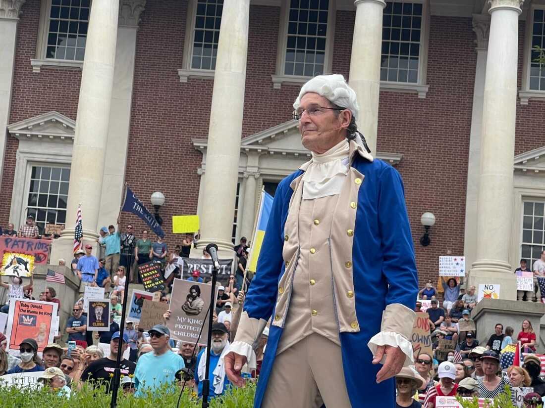 乔治·华盛顿（George Washington）的重演者在一座红砖建筑前戴着白色假发和蓝色背心时发表演讲。