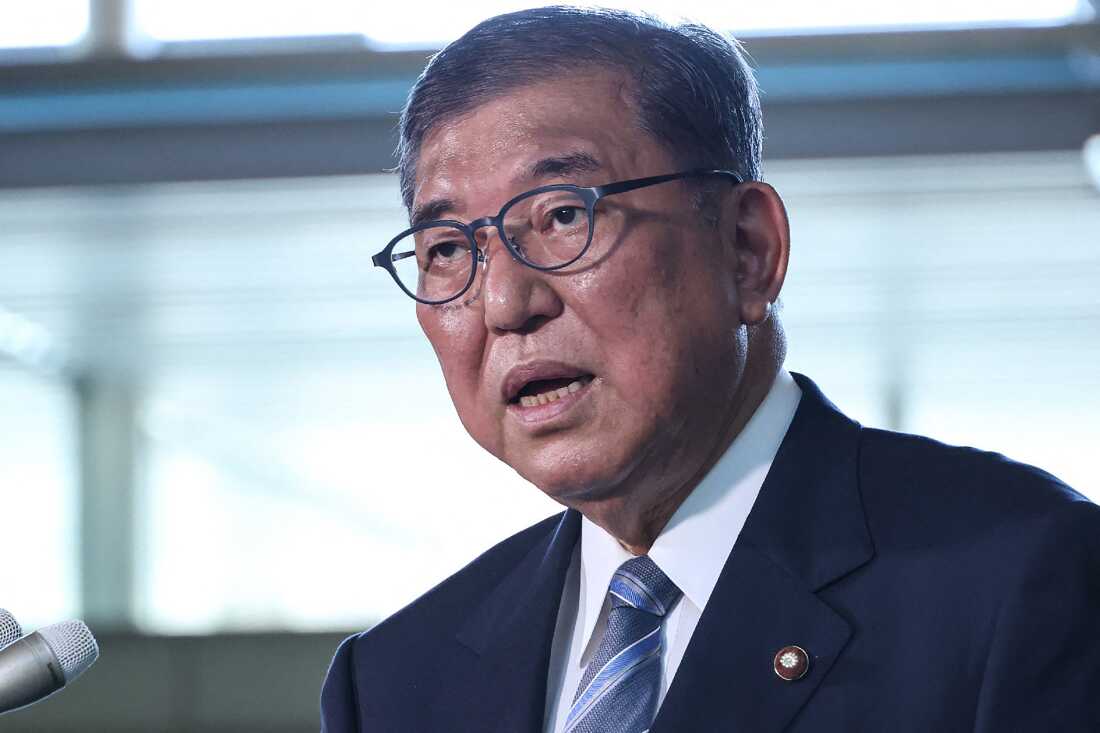 这张照片显示，日本总理Shigeru Ishiba从肩膀上拍照。他穿着西装和眼镜，正在用麦克风说话。