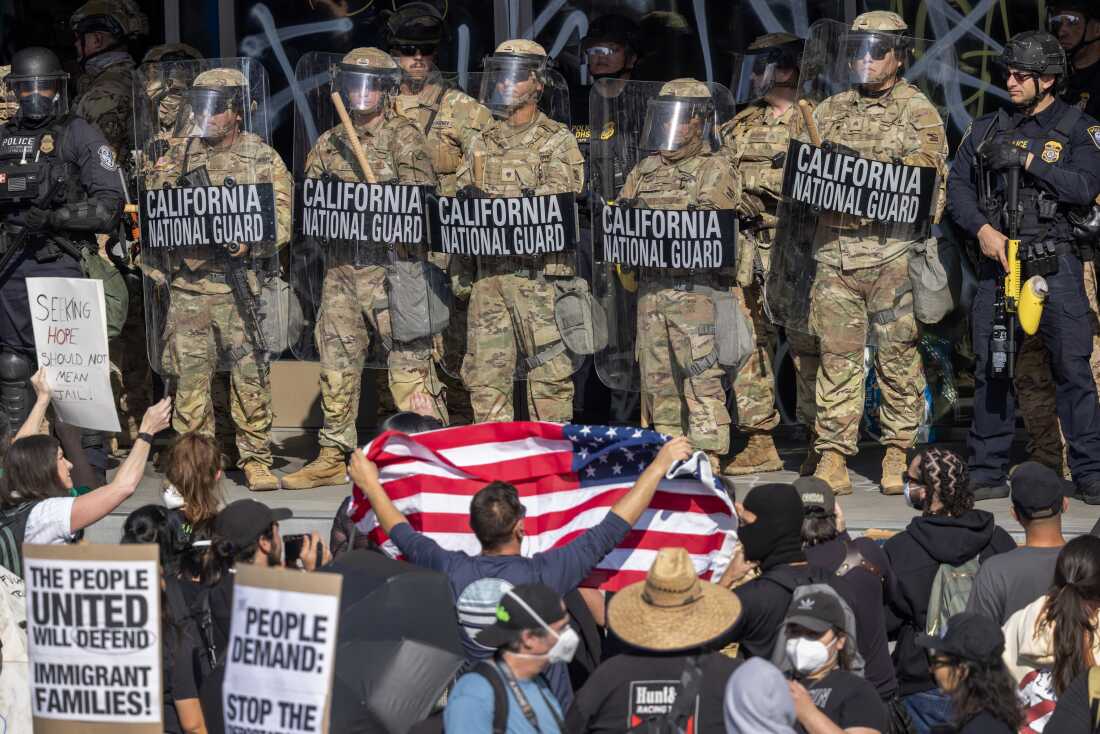 加利福尼亚国民警卫队士兵和警察于2025年6月连续站在洛杉矶。国民警卫队士兵穿着伪装制服，并带有透明的盾牌。在前台立场抗议者中，其中一些人带有迹象。