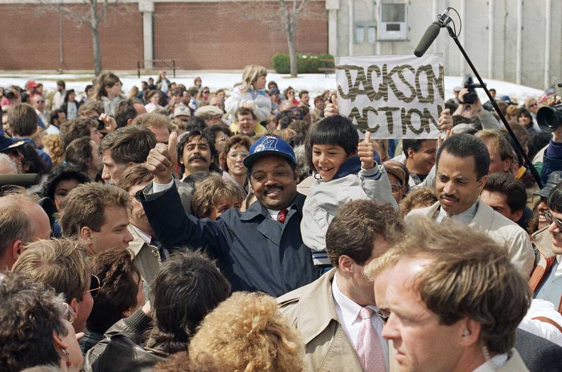 1988 年 4 月 2 日，科罗拉多州格里利，民主党总统候选人杰西·杰克逊牧师和 8 岁的拉里·埃斯托达在杰克逊竞选总统集会上竖起大拇指。杰克逊在一大群人中间抱着埃斯托达。他们身后有一个手绘的牌子，上面写着 "杰克逊行动。"