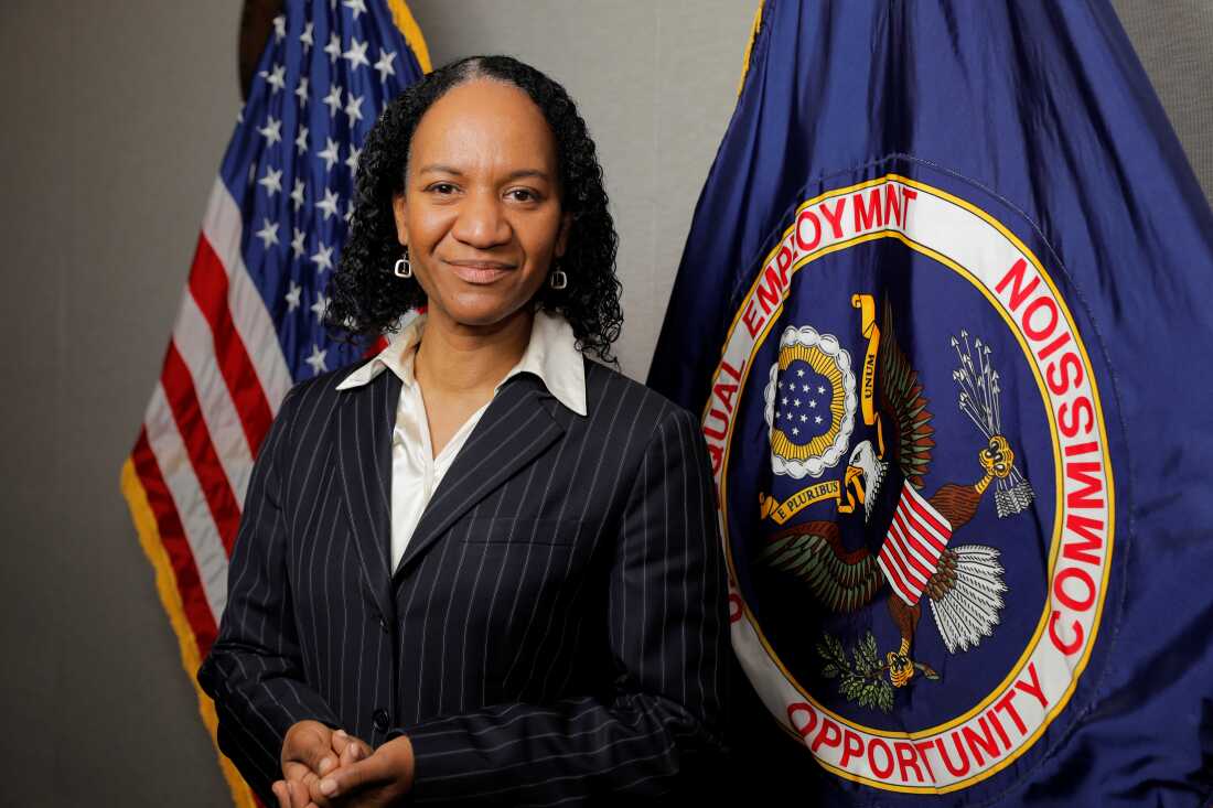2021 年，当时的 EEOC 主席夏洛特·伯罗斯 (Charlotte Burrows)。她是一名黑人女性，站在美国国旗和 EEOC 旗帜前，旗帜是蓝色的，上面有一只鹰。