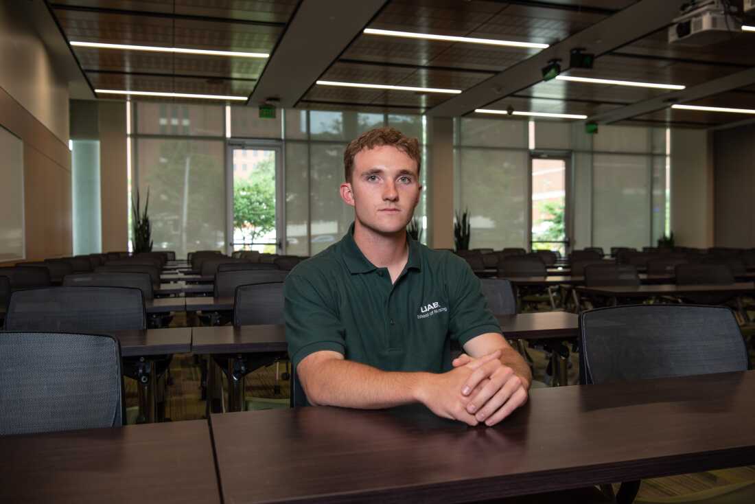 护理学生 Connor Aderholt 留着短卷发，穿着 UAB Polo 衫。他坐在教室的一张桌子旁。